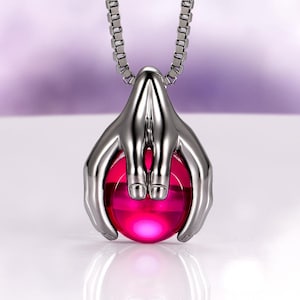 May include: A silver-toned pendant necklace featuring a stylized pair of hands cradling a vibrant pink orb. The necklace has a chain with square links. The pendant's design is modern and eye-catching, suitable for everyday wear.