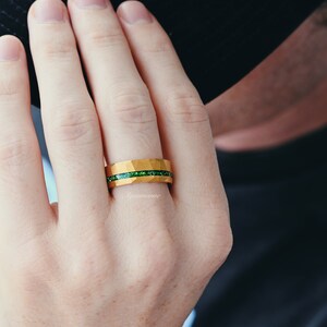 K&ouml;nnte beinhalten: Ein goldener Ring mit einem gr&uuml;nen Edelsteinband. Der Ring befindet sich an einem Finger.