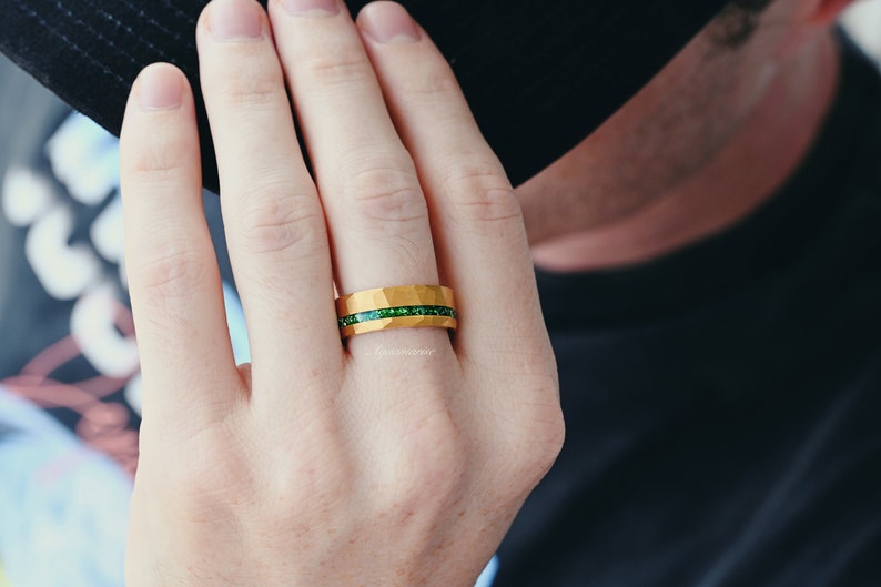 K&ouml;nnte beinhalten: Ein goldener Ring mit einem gr&uuml;nen Edelsteinband. Der Ring befindet sich an einem Finger.