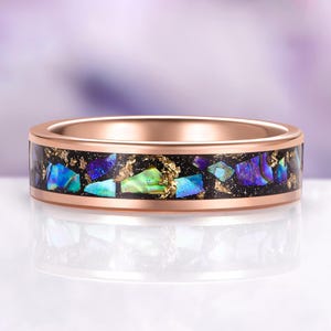May include: Rose gold ring with a black inlay featuring iridescent blue, green, and purple abalone shell fragments and gold flakes. The ring has a polished finish and a smooth, rounded band. A unique piece of jewelry.