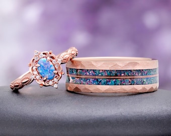 May include: A rose gold engagement ring featuring an oval blue opal center stone surrounded by small clear accent stones, paired with a wide rose gold wedding band with a hammered finish and a central inlay of crushed blue and green opal flakes.