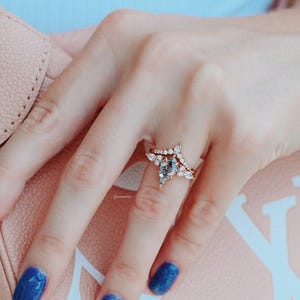 May include: A close-up of a hand wearing a rose gold ring with a pear-shaped center stone and a delicate band of smaller stones. The ring is set on a finger with blue nail polish.