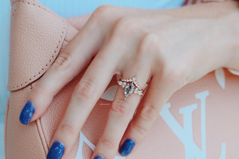 May include: A close-up of a hand wearing a rose gold ring with a pear-shaped center stone and a delicate band of smaller stones. The ring is set on a finger with blue nail polish.
