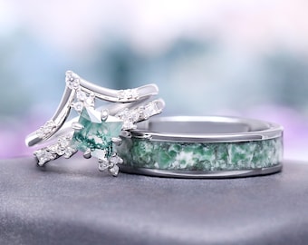 May include: Two rings, one with a square green gemstone and small clear stones, the other with a band of green and white stone. Both rings are silver in color and sit on a gray surface.