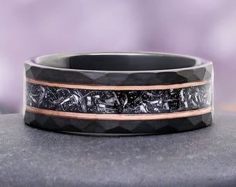 May include: A black ring with a textured surface and a copper-colored band. The center band features a dark, patterned design. The ring is displayed on a dark gray surface against a blurred purple background.