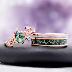 May include: A rose gold ring featuring an oval moss agate stone and small opal accents alongside a rose gold band inlaid with a horizontal strip of white opal above a wider band of dark green moss agate with a polished metallic finish.