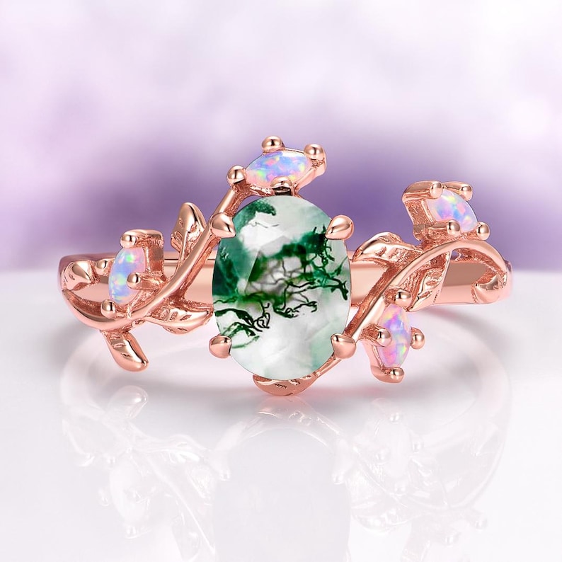 May include: A rose gold ring featuring a central oval moss agate stone with green inclusions, flanked by leaf-shaped metal accents and four small, iridescent, oval-shaped opals set along the band in a vine-like arrangement.