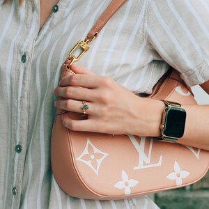 May include: A gold ring featuring a central oval green gemstone and small clear accent stones, worn on a finger. The ring is displayed alongside a peach-coloured shoulder bag with a white monogram pattern and a gold-tone smartwatch with a mesh band.
