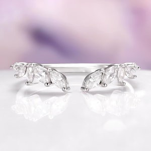 May include: A silver-toned ring with a unique design. The ring features a series of marquise-cut clear gemstones arranged in a curved, open design. The gemstones are set closely together, creating a sparkling effect. The ring is set against a soft, blurred background.