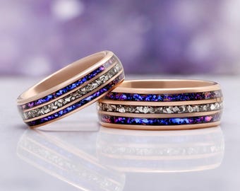 May include: Two rose gold rings featuring a central inlay of crushed silver-toned metallic flakes flanked by two bands of iridescent blue and purple crushed material. The rings have a smooth, polished finish and a rounded profile design.