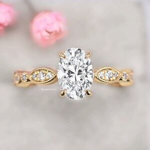 Puede incluir: Un anillo de compromiso de oro con una piedra central de diamante de forma ovalada y una banda delicada e intrincada adornada con diamantes m&aacute;s peque&ntilde;os.