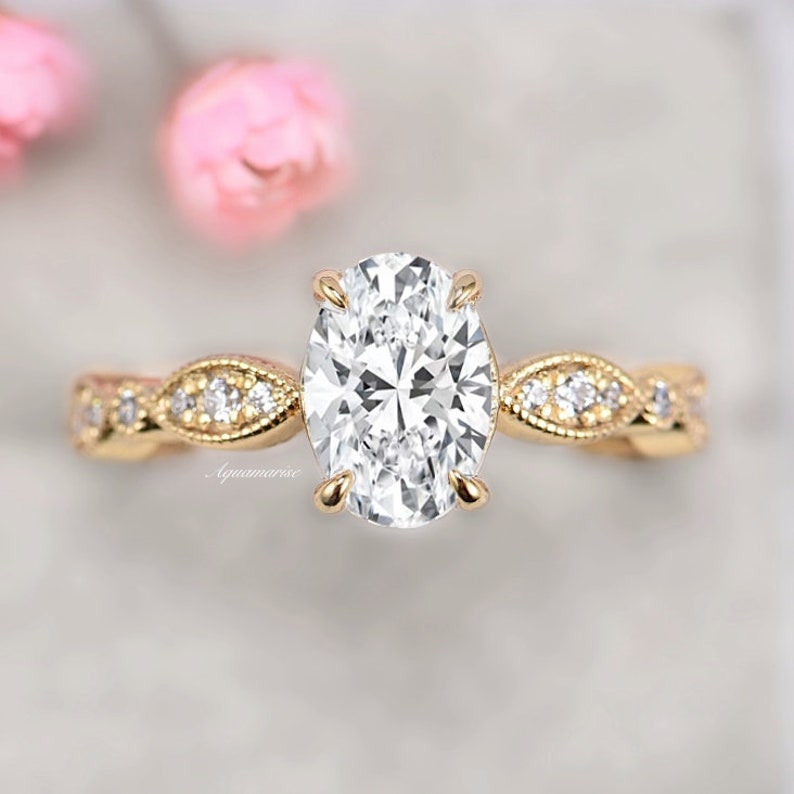 Puede incluir: Un anillo de compromiso de oro con una piedra central de diamante de forma ovalada y una banda delicada e intrincada adornada con diamantes m&aacute;s peque&ntilde;os.