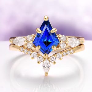 May include: A gold engagement ring set featuring a kite-shaped, deep blue gemstone as the centerpiece. The ring is accented with smaller, clear gemstones and a matching band, creating a sparkling and elegant design.