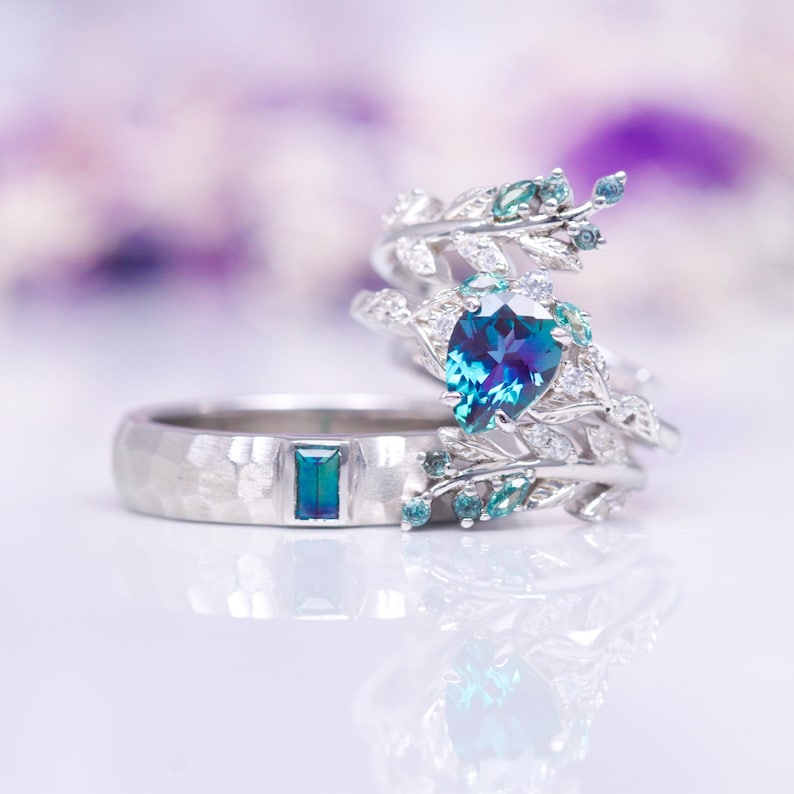 Op de afbeelding: Een paar zilveren trouwringen met blauwe en groene edelstenen. De verlovingsring heeft een peervormige blauwe edelsteen omringd door kleinere diamanten en groene edelstenen in een bladontwerp. De trouwring heeft een gehammerde afwerking en een rechthoekige blauwe edelsteen.