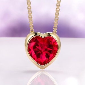 May include: A gold-colored necklace with a heart-shaped, ruby-red gemstone pendant. The pendant is encased in a gold bezel and hangs from a delicate gold chain. The gemstone has multiple facets, reflecting light.