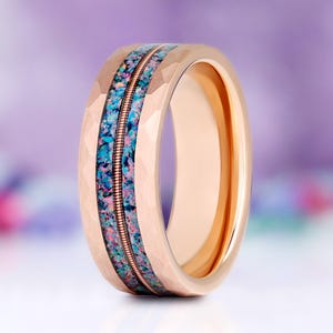 May include: A rose gold-toned ring with a faceted outer surface and a central inlay. The inlay features a vibrant mix of crushed blue and purple stone fragments bisected by a thin, metallic guitar string element running horizontally.