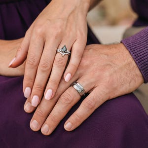 Pu&ograve; includere: Un set di anelli di fidanzamento color argento con una pietra centrale grigio scuro a forma di aquilone e piccole pietre bianche, abbinato a una fede nuziale larga e sfaccettata con un canale centrale incastonato di piccole pietre chiare.