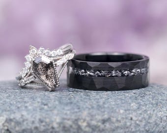 Może przedstawiać: Para obrączek ślubnych. Pierścionek zaręczynowy ma duży, szary, diamentowy kamień otoczony mniejszymi diamentami. Obrączka ślubna jest czarna z młotkowanym wykończeniem i rzędem małych diamentów.