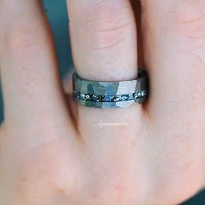 Pu&ograve; includere: Un anello in metallo martellato grigio scuro caratterizzato da un canale orizzontale centrale riempito con piccole scaglie scintillanti blu e argento. La parola Aquamarise &egrave; stampata in un delicato carattere corsivo bianco sotto l'anello.