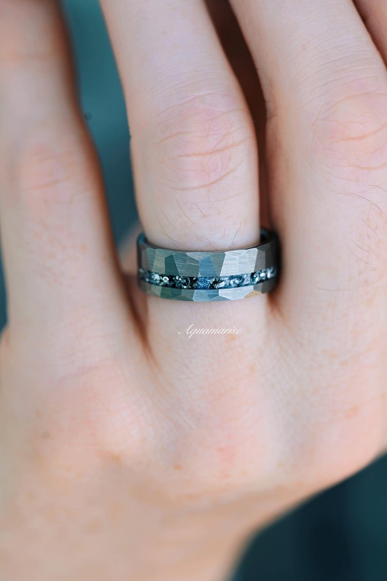 Pu&ograve; includere: Un anello in metallo martellato grigio scuro caratterizzato da un canale orizzontale centrale riempito con piccole scaglie scintillanti blu e argento. La parola Aquamarise &egrave; stampata in un delicato carattere corsivo bianco sotto l'anello.