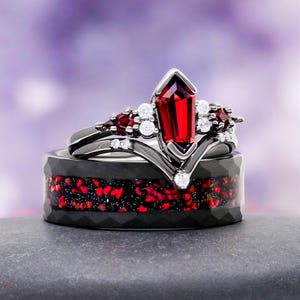 May include: A set of two rings. The top ring is black with a large, red gemstone and smaller white and red stones. The bottom ring is black with a red and black speckled inlay. The rings are displayed on a gray surface.