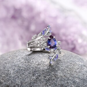 Op de afbeelding: Een ring met een peer-vormige paarse edelsteen, een zilveren band en delicate bladvormige details die zijn versierd met kleinere paarse edelstenen.