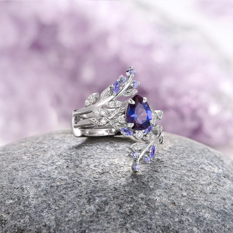 Op de afbeelding: Een ring met een peer-vormige paarse edelsteen, een zilveren band en delicate bladvormige details die zijn versierd met kleinere paarse edelstenen.