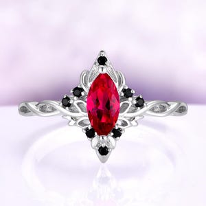 May include: A silver ring featuring a marquise-cut, vibrant red gemstone at the center, surrounded by black accent stones. The ring has an ornate, floral-inspired design with a twisted band.
