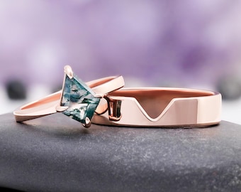 May include: Two rose gold rings. One ring features a square-cut, moss agate gemstone with green and blue inclusions. The other ring has a unique design with a V-shaped cutout. Both rings are displayed on a gray stone.