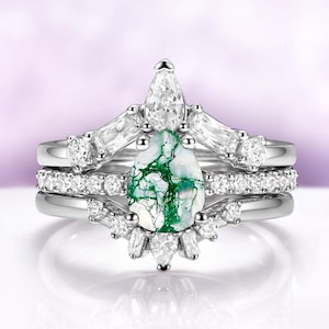 May include: A three-piece ring set featuring a pear-shaped moss agate gemstone with green veining. The set includes a main ring and two bands, all with silver-colored metal and clear accent stones. The main ring has a crown-like design.