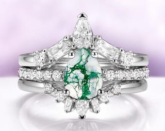 May include: A three-piece ring set featuring a pear-shaped moss agate gemstone with green veining. The set includes a main ring and two bands, all with silver-colored metal and clear accent stones. The main ring has a crown-like design.