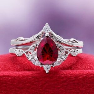 May include: A silver-toned ring set featuring a central pear-shaped deep red gemstone. The band and setting are decorated with intricate leaf-like metalwork and small, clear, round-cut accent stones arranged around the primary red stone.