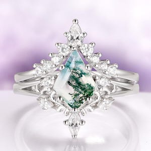 May include: A silver engagement ring set with a unique, geometric moss agate center stone. The ring is surrounded by small, sparkling diamonds and features a matching band. The moss agate has green and white patterns.