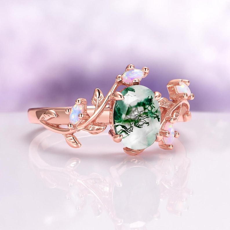 May include: A rose gold ring featuring a central oval moss agate gemstone with green inclusions. The band is designed with metal vine and leaf motifs, accented by four small, iridescent marquise-cut opals set along the branches.