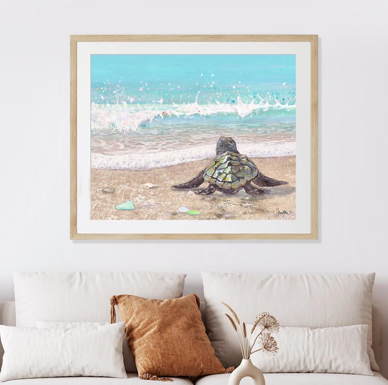May include: A framed watercolor painting depicts a sea turtle on a sandy beach, facing the ocean. The artwork features a turquoise sky and ocean with white waves. The turtle has a green and brown shell. The painting is in a light wood frame.