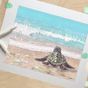 May include: A framed print of a sea turtle on a sandy beach, with a turquoise ocean and white waves in the background. The artwork is surrounded by a white mat, with a starfish and sea glass accents.