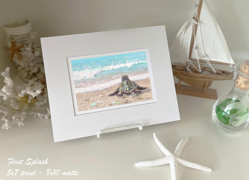 May include: A framed print of a sea turtle on a sandy beach, facing the ocean waves. The artwork is displayed in a white mat and frame. Decorative elements include a small sailboat, starfish, and a glass jar.