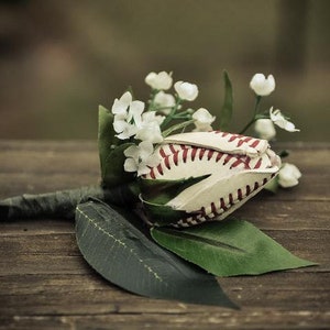 May include: A unique baseball-themed floral arrangement. The centerpiece is a baseball, with red stitching, transformed into a flower. White flowers and green leaves are arranged around the baseball, creating a rustic, sporty aesthetic.