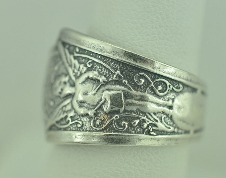 May include: Silver ring with an intricate design featuring a winged figure and floral motifs.