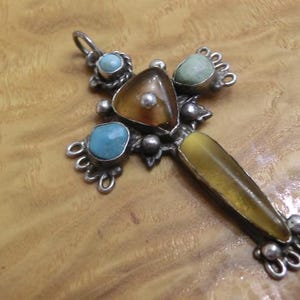 Pode incluir: Um pingente de cruz prateado com pedras preciosas coloridas. A cruz apresenta uma pedra central marrom, rodeada por pedras turquesas e verdes. O braço da cruz tem uma pedra amarelo-castanho. O pingente está sobre uma superfície de madeira.