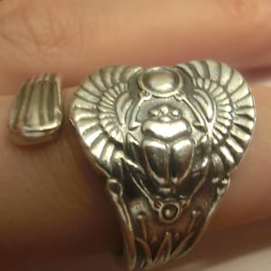 May include: A silver-toned ring featuring a central scarab beetle motif with outstretched wings. The design includes a circular element above the beetle and stylized floral patterns along the band. The ring has an open, adjustable-style construction.