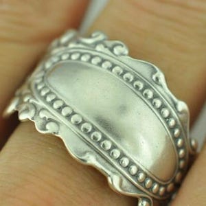May include: A wide silver ring featuring a smooth, polished central oval panel framed by a raised border of small, uniform beads. The edges of the band are scalloped with decorative, curved metalwork that extends along the entire outer perimeter.