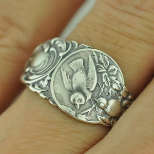 May include: A silver ring featuring an embossed design of two birds perched on a branch surrounded by intricate floral scrollwork. The metal has a polished finish with darkened recessed areas to highlight the detailed relief of the avian motif.