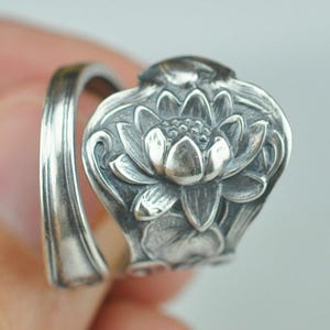 May include: A silver-toned spoon ring featuring a detailed, embossed lotus flower design on the face. The band is formed from the handle of a spoon, showing a smooth, curved metal surface that wraps around to meet the floral centerpiece.