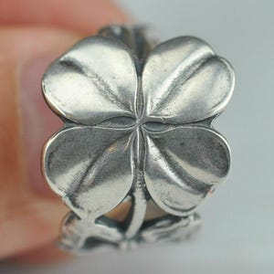 Può includere: Un anello color argento caratterizzato da un prominente design a quadrifoglio sulla parte anteriore. Il quadrifoglio presenta petali lisci e arrotondati con venature definite e una finitura metallica lucida. La fascia dell'anello prosegue il motivo.