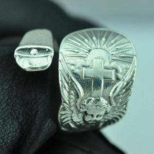 May include: A silver-toned wrap ring featuring an embossed design of a cherub with wings, a central cross, and a sunburst pattern above. The band is open-ended with a smooth, rounded terminal on one side and a wide, detailed face on the other.