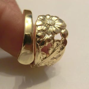 May include: A gold-colored ring featuring a wide band with an openwork design of a daisy flower and leaves. The band has a smooth, polished finish on one side and a detailed, raised floral pattern on the other, creating a wrap-around appearance.