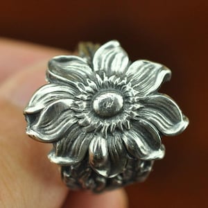 May include: A silver ring featuring a detailed, dimensional flower design on the face. The petals are sculpted with deep grooves and raised edges, radiating from a central circular disc. The metal has a polished finish with darkened accents.