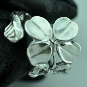 Può includere: Un anello color argento caratterizzato da un design a quadrifoglio come punto focale centrale. La fascia dell'anello ha la forma di uno stelo sottile e curvo che avvolge il dito, collegandosi alle foglie con una finitura lucida.