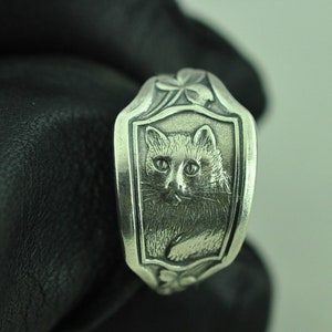 May include: A silver ring with a detailed design of a cat's face in a shield-shaped frame. The ring is decorated with clover leaves.